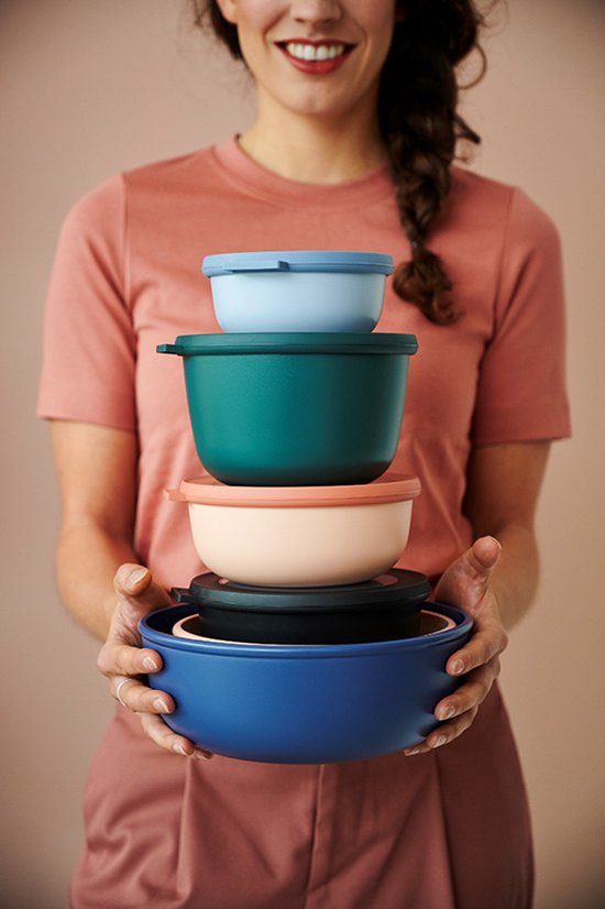 Une femme avec une tresse, vêtue d'un t-shirt rose, tient une pile de cinq boîtes de conservation colorées. De bas en haut : bleu, vert, rose pâle, noir et bleu clair.