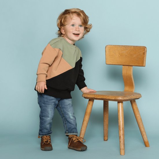 Jongen met blonde krullen, staand naast een houten stoel, draagt een bruin, beige en zwart colorblock sweater en blauwe spijkerbroek.