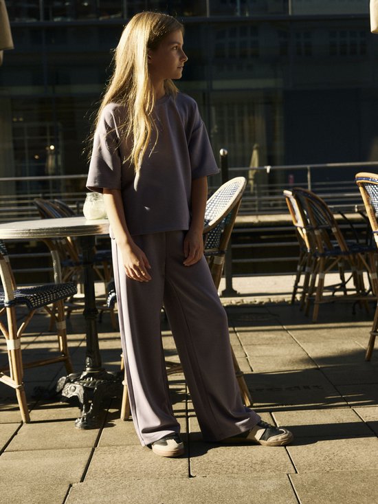 Meisje draagt een grijze set met een T-shirt met korte mouwen en een broek met wijde pijpen op een terras.