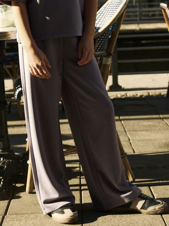 Detail van een meisje in een grijze broek met wijde pijpen en sneakers op een terras.
