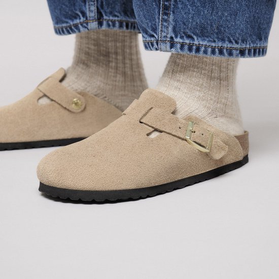 Persoon draagt sandkleurige suède Birkenstock clogs met gebreide sokken en spijkerbroek.