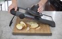 Een mandoline snijdt appelpartjes op een houten snijplank. Een hand houdt de voedselhouder stevig vast.