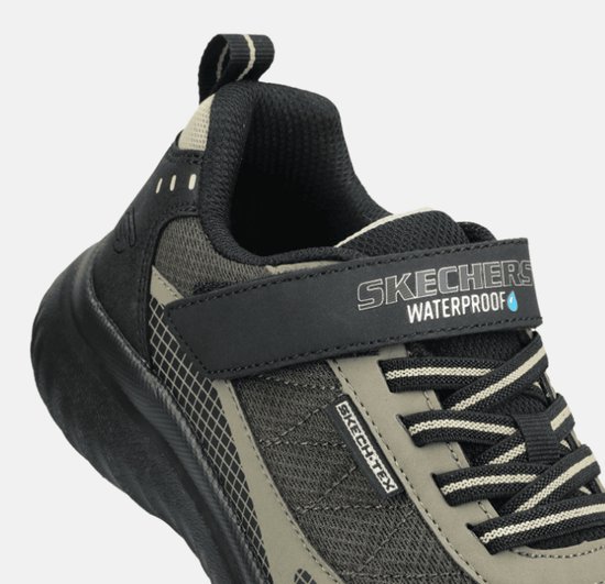 Detail van zwarte sneaker met klittenband met 'Skechers Waterproof' tekst. Grijze en beige mesh.