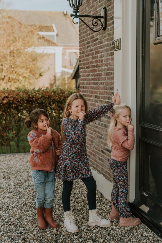 Drie meisjes staan bij een deur. Twee dragen een roze jasje met een gebloemde jurk, en een met een bruine teddy jas en spijkerbroek. Beide jongste meisjes dragen dezelfde flared broek.