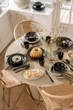 Gedekte ronde tafel met zwart servies, gebak en rieten stoelen.