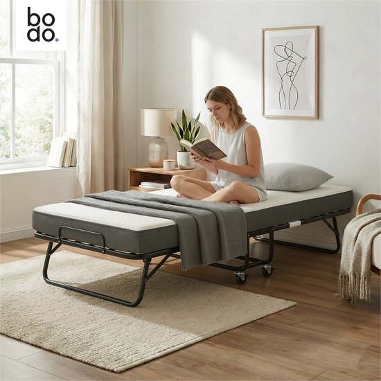 Vouwbed van Bodo met donkergrijs matras, uitgeklapt met een vrouw die leest. Het bed heeft een zwart metalen frame en wieltjes.