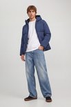 Man met donkerblauwe pufferjas met capuchon, witte t-shirt en wijde spijkerbroek.