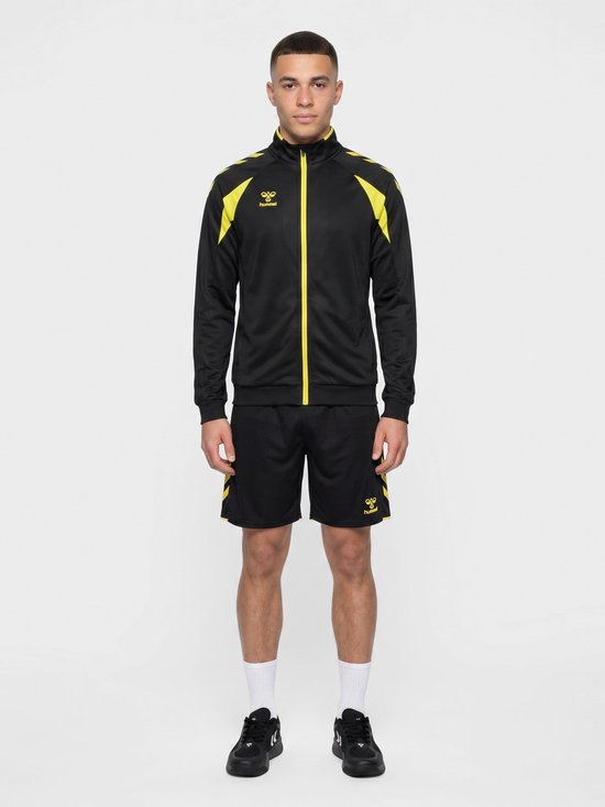 Man draagt zwarte Hummel trainingsjas met gele accenten en bijpassende shorts, van voren gezien.