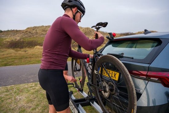 Man in donkerwijnrode trui bevestigt een racefiets op een trekhaakfietsendrager achter een auto.