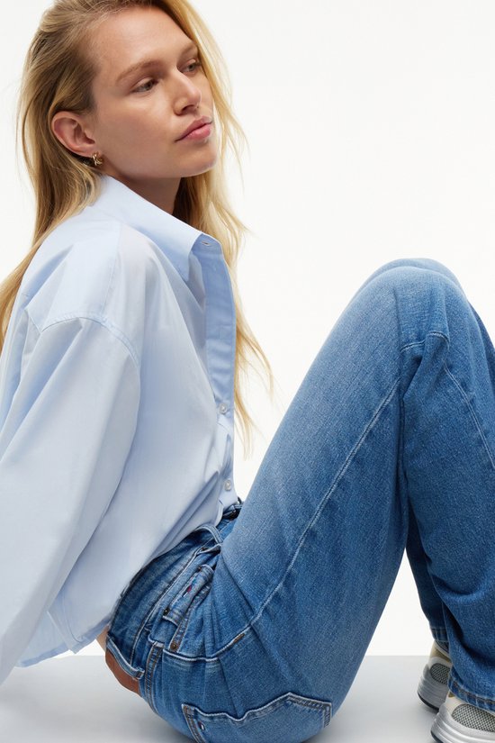 Model zittend in een lichtblauwe oversized blouse en spijkerbroek, close-up van de taille.