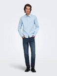 Man in lichtblauwe blouse met lange mouwen en donkere jeans, handen in de zakken.