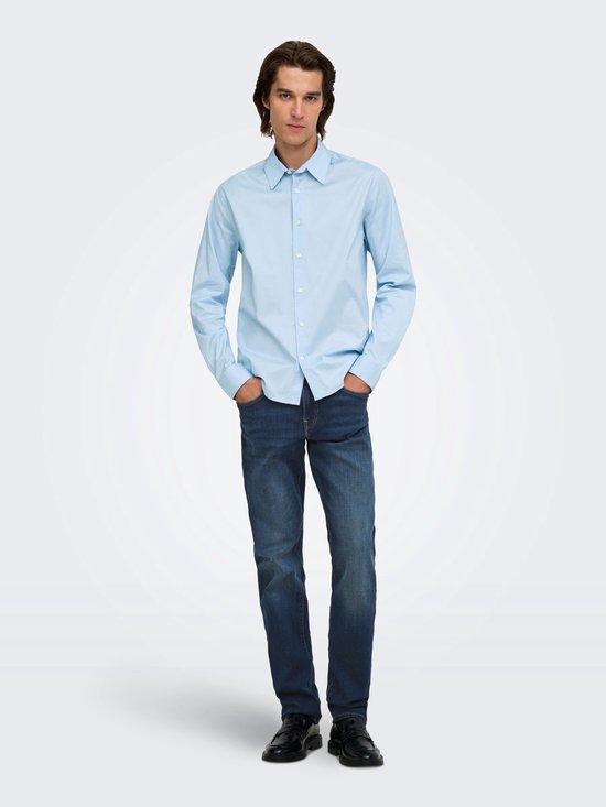 Man in lichtblauwe blouse met lange mouwen en donkere jeans, handen in de zakken.