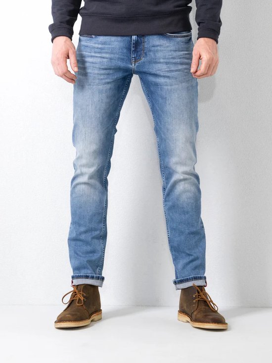 Man in een blauwe jeans met opgerolde broekspijpen en bruine schoenen. De jeans heeft een lichte wassing.