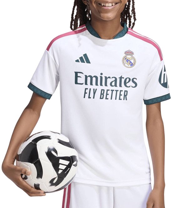 Een kind in een wit voetbalshirt met roze strepen op de schouders en groen aan de zomen, met een voetbal in de hand. De tekst 'Emirates FLY BETTER' is zichtbaar.