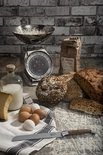 Brood, kaas, eieren, melk, bloem en een broodmes met olijfhouten handvat op een houten ondergrond.