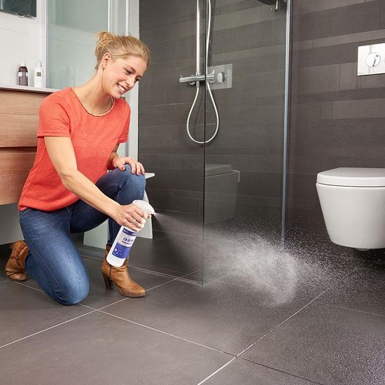 Vrouw met oranje shirt en spijkerbroek knielt en spuit anti-slip spray op donkergrijze vloertegels bij douche.