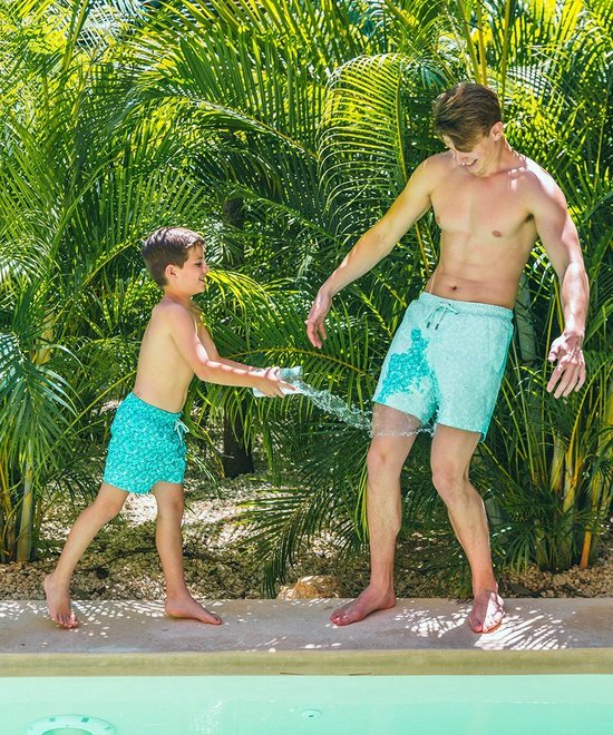 Man en jongen met zwembroeken in het water, omringd door palmbomen.