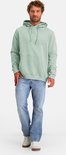 Staande man in mintgroene hoodie met logo en jeans op witte achtergrond.