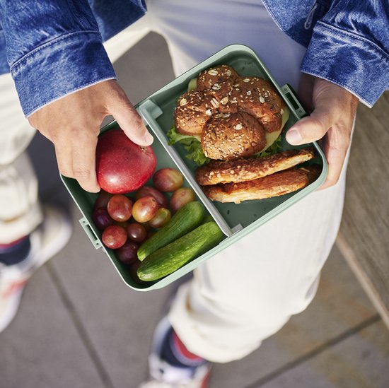 Personne portant une veste en jean ouvre une boîte à lunch verte contenant des petits pains, du blanc de poulet, du concombre, des raisins et une pomme.
