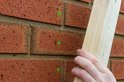 Een hand houdt een houten plank tegen een rode bakstenen muur met groene markeringen.