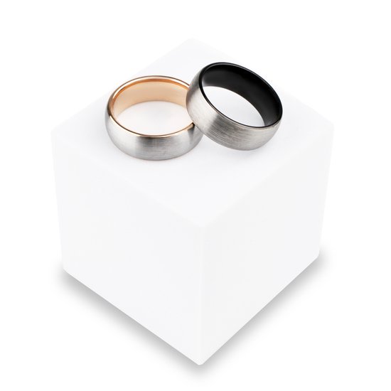 Twee ringen op een witte kubus: één met rosé gouden binnenkant, één met zwarte binnenkant en stalen buitenkant.