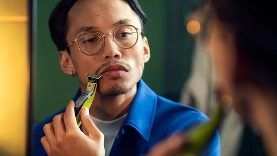Man met bril trimt zijn snor en baard met een groene en zwarte Philips OneBlade, kijkend in de spiegel.