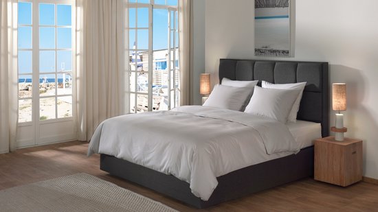 Een opgemaakt bed met Vaporous Grey dekbedovertrek, bij een raam met uitzicht op zee.