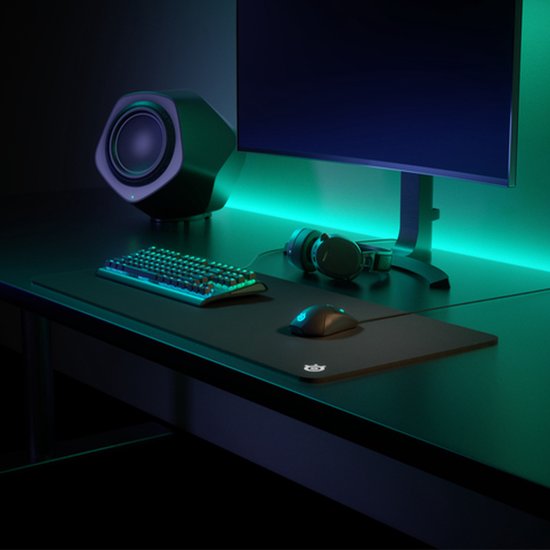 Zwarte gaming muismat met SteelSeries logo, met daarop een muis en toetsenbord met RGB-verlichting. Een monitor en koptelefoon staan ernaast, verlicht door groen en paars licht.