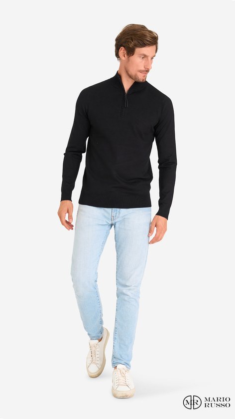 Man met een zwarte trui met rits en lichtblauwe jeans, kijkend naar de zijkant.