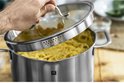 Een hand tilt het glazen deksel van een RVS pan met spaghetti op. De deksel heeft geperforeerde randen.