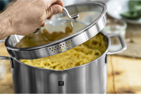 Een hand tilt het glazen deksel van een RVS pan met spaghetti op. De deksel heeft geperforeerde randen.
