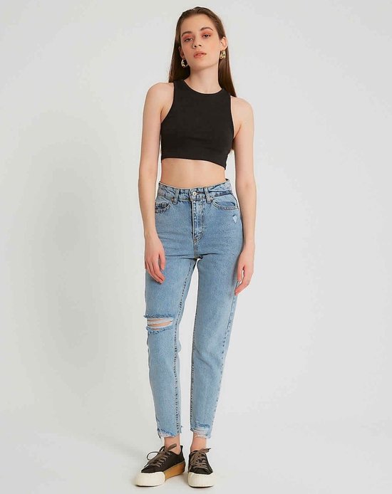 Model draagt een lichtblauwe spijkerbroek met hoge taille en een gescheurde knie. Ze draagt ook een zwarte crop top en sneakers.