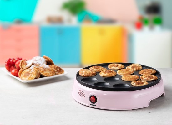 Roze poffertjespan op een tafel met op de achtergrond een kleurrijke keuken en een bord poffertjes.