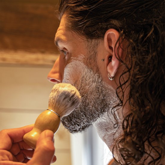 Man met baard en scheerschuim gebruikt een scheerkwast met bamboe handvat en bruin-witte haren.