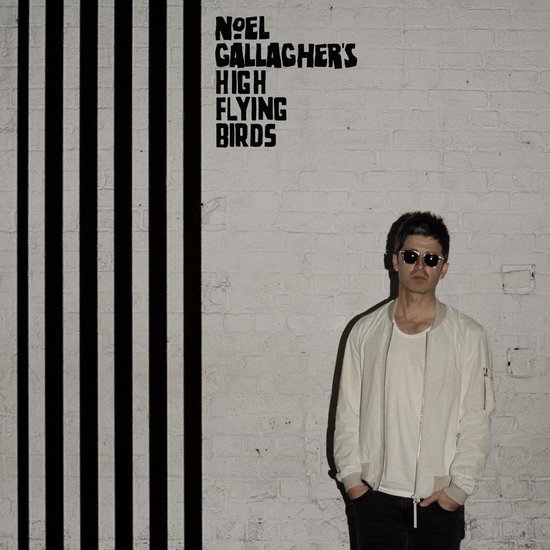 Hoes met een man met zonnebril en een wit jack, tegen een bakstenen muur met zwarte strepen. De tekst 'NOEL GALLAGHER'S HIGH FLYING BIRDS' staat erboven.
