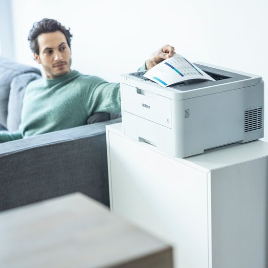 Man pakt een document uit een witte Brother printer op een witte kast.