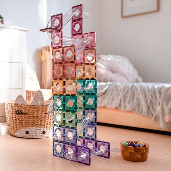 Een hoge toren van magnetische Connetix-tegels in pastelkleuren, met transparante buizen, op een roze vloer in een kinderkamer.