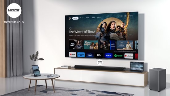 Zwarte soundbar en subwoofer naast een tv die op een laag meubel staat. Een laptop en tablet zijn ook zichtbaar.