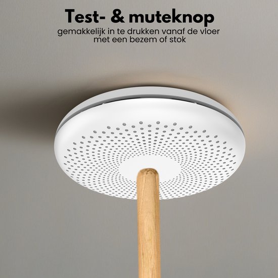 Witte, ronde rookmelder met cirkelvormig patroon van gaatjes, gemonteerd aan het plafond. Een houten stok raakt het midden.