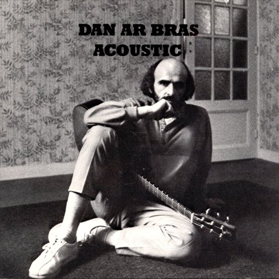 Dan Ar Bras zit met een akoestische gitaar op schoot, gekleed in een trui en broek.
