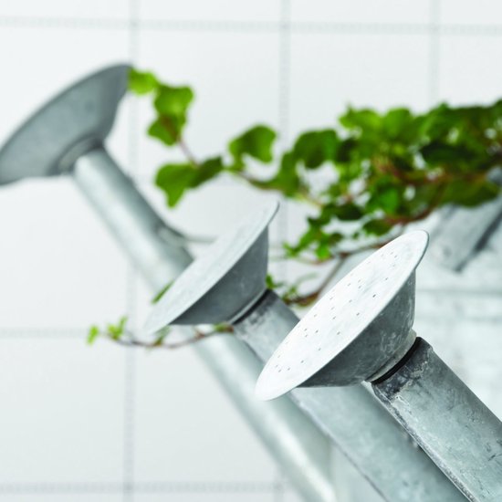 Drie metalen gieters met groene plantentakken op de voorgrond, witte tegels op de achtergrond.