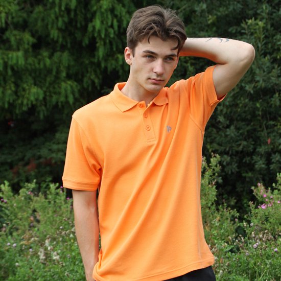 Model dat in de natuur staat, met een hand in de zak, in een oranje polo met korte mouwen.