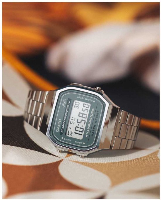 Zilverkleurig Casio horloge met groen digitaal display op een stoffen ondergrond met bloemenpatroon.