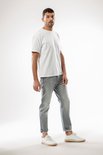 Man in witte t-shirt en grijze jeans, staand met handen in de zakken.