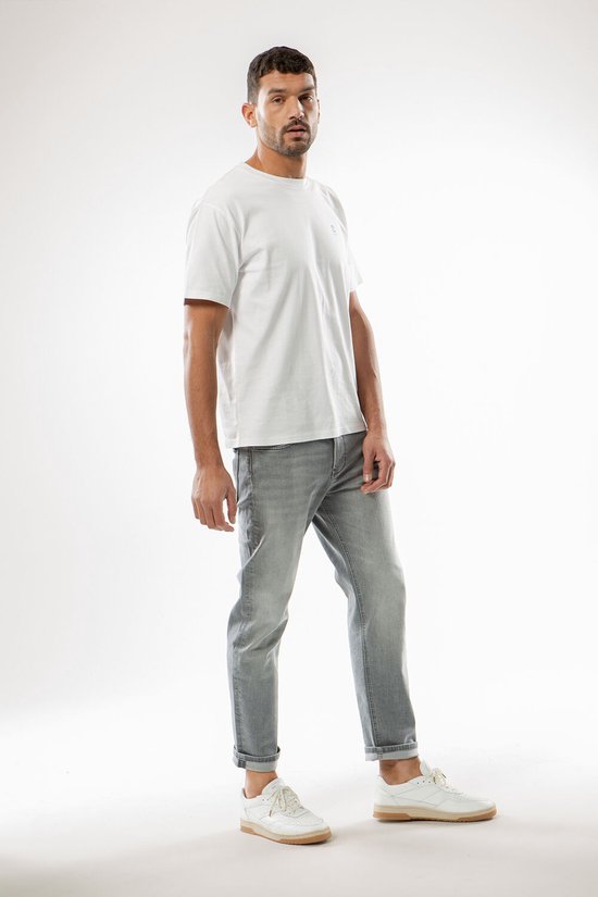 Man in witte t-shirt en grijze jeans, staand met handen in de zakken.