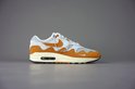 Linker Nike Air Max 1 Patta sneaker in wit, oranje, beige en bruin, op een grijze achtergrond.