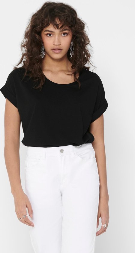 Vrouw draagt een zwarte top met opgerolde korte mouwen en een witte jeans. Ze draagt opvallende oorbellen.