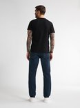Man met tatoeages op armen, gekleed in een zwarte t-shirt en een donkerblauwe jeans.