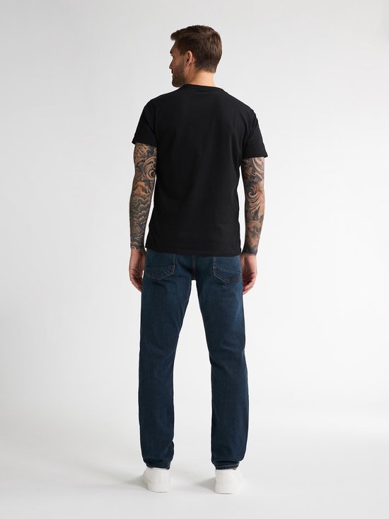 Man met tatoeages op armen, gekleed in een zwarte t-shirt en een donkerblauwe jeans.
