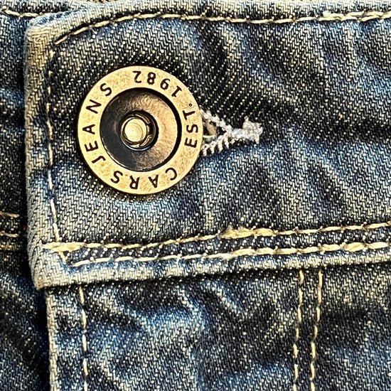 Detailopname van de metalen knoop van de spijkerbroek met de tekst 'CARS JEANS EST. 1982'.